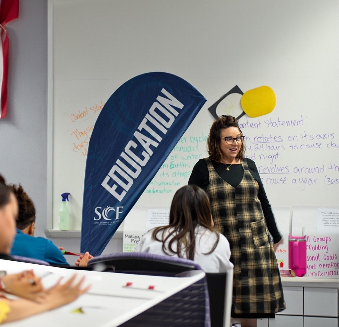 Teacher stands at the whiteboard at the front of a classroom with "education" banner